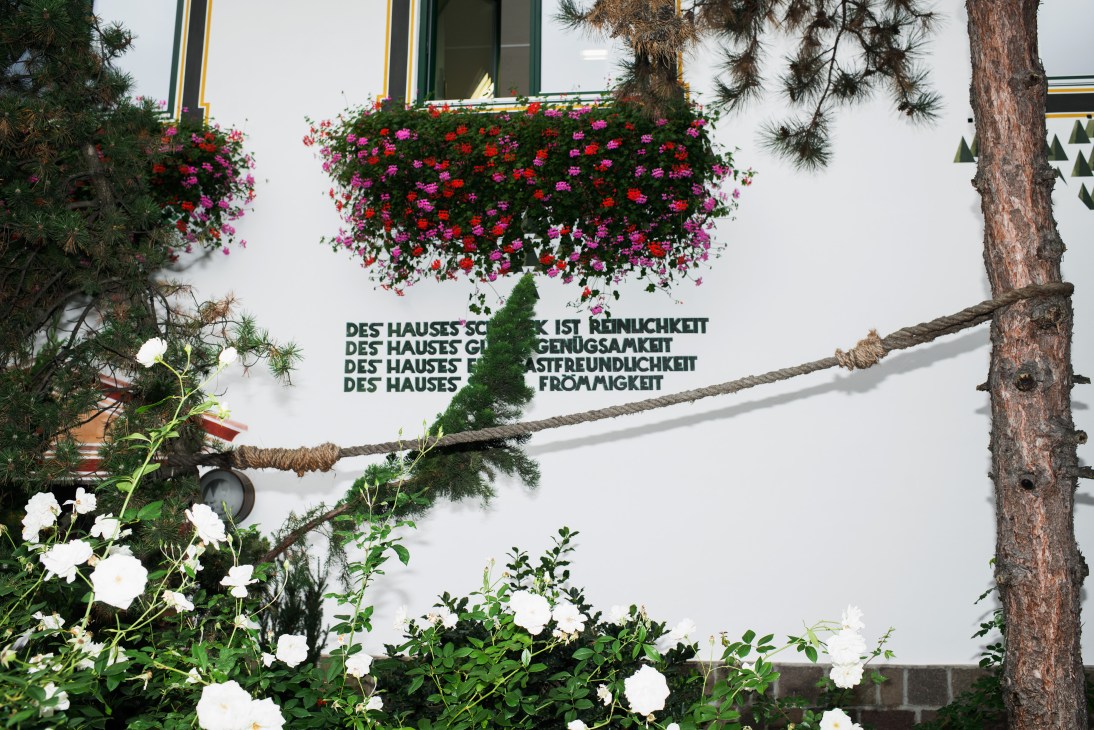 Hanging flower baskets with pink and red blooms above German text on brewery wall.