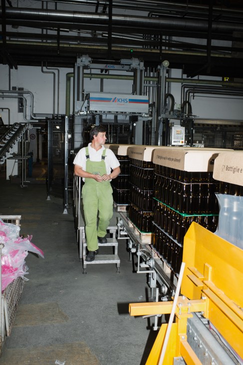 Technician in green work overalls stands in Forst brewery's bottling plant amid industrial machinery and conveyor systems.