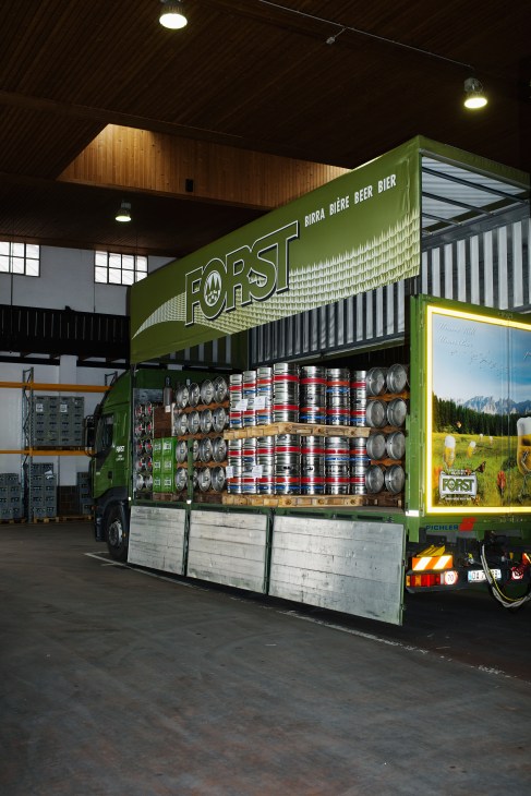 A green Forst delivery truck loaded with beer kegs and cans inside a warehouse.
