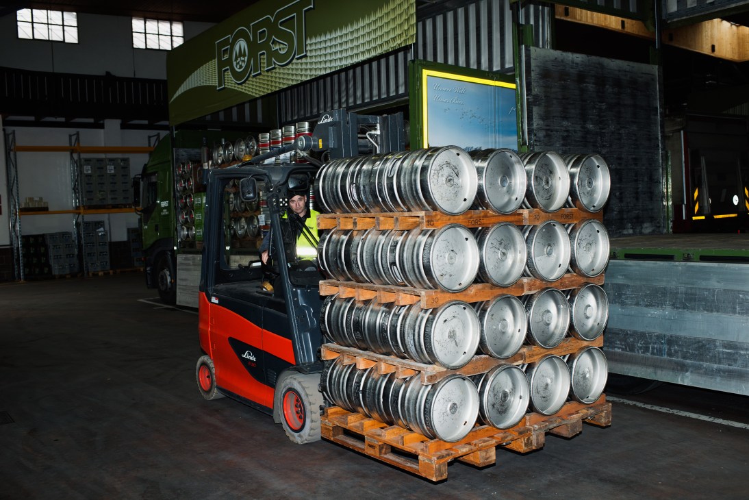Forst brewery worker operating red and black Linde forklift loaded with stacked metal beer kegs on wooden pallets.