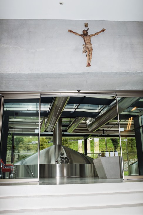 Wooden crucifix mounted on white wall above modern glass brewery entrance with stainless steel equipment visible inside.