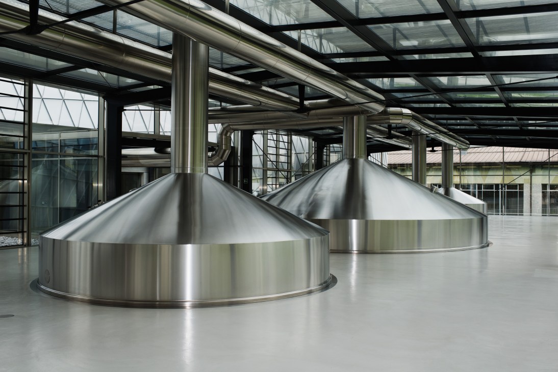 Two large stainless steel conical brewing vats with peaked metal hoods inside a modern industrial brewery facility with steel support beams and glass roof.