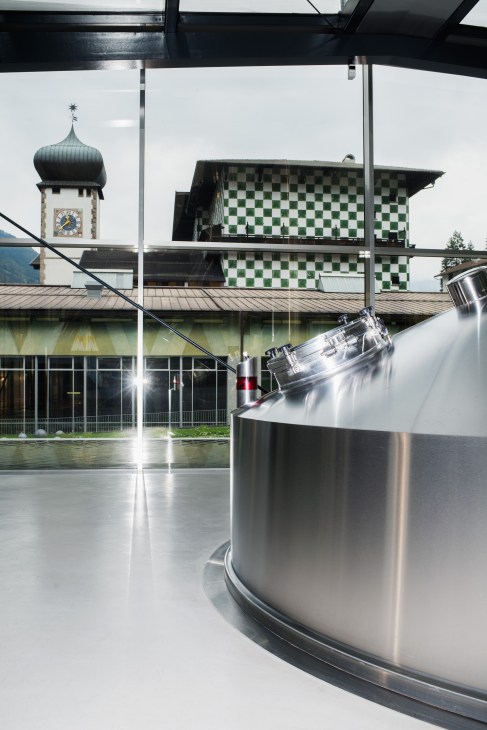 Large stainless steel mash tun at Forst brewery with Alpine bell tower and checkered building visible in background.