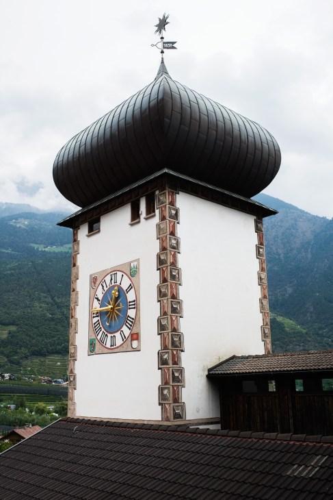 Forst brewery's distinctive belltower with black onion dome and 1857 weathervane against Alpine mountain backdrop.