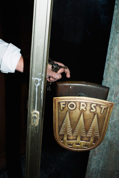 Hand holding a key next to a brass Forst brewery logo featuring three alpine trees on a door.