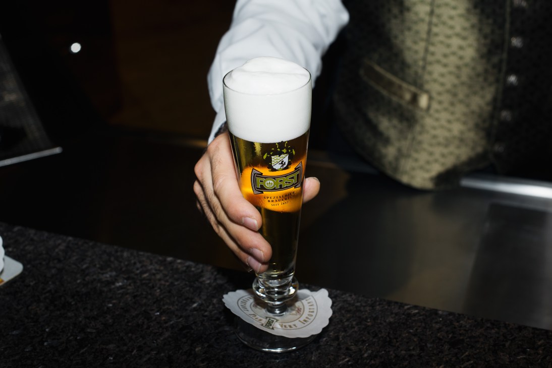 Hand holding a glass of Forst VIP pilsener beer with foam head and logo visible on the glass.