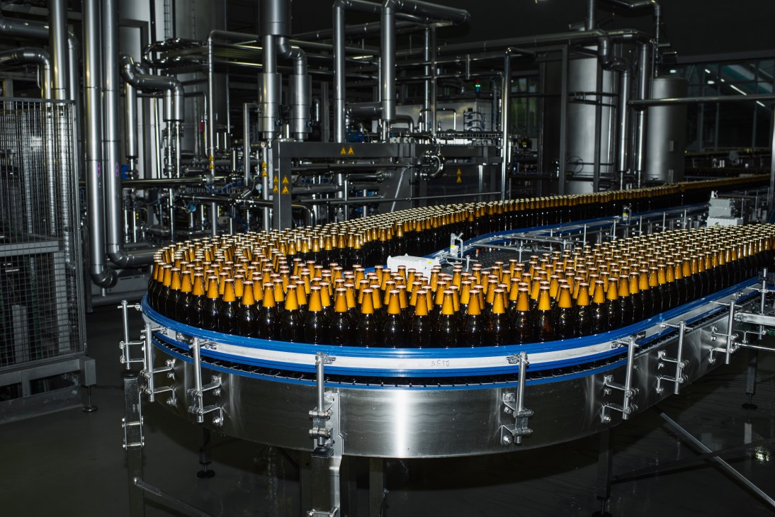 Forst beer bottles on a blue conveyor belt in the brewery's bottling plant with stainless steel machinery in the background.
