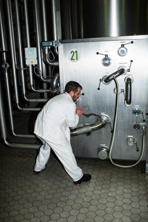 A technician in white work clothes operates control valves and gauges on stainless steel brewery equipment.