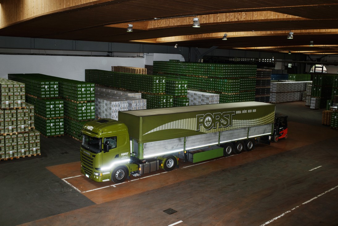 A bright lime-green Forst beer delivery truck in a warehouse surrounded by stacked crates of beer bottles.