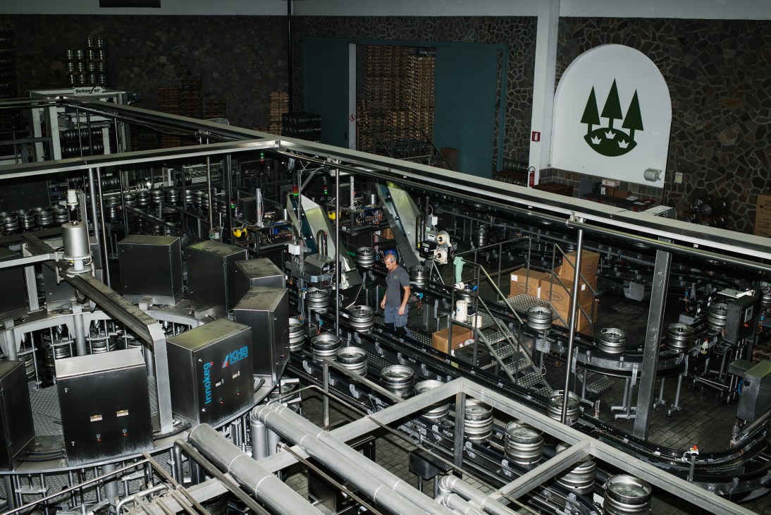 Worker in Forst brewery's kegging plant with metal kegs and machinery, displaying the three-tree logo on wall.