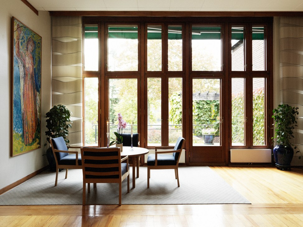 Mid-century modern lounge with teak furniture, colorful artwork, and expansive teak-framed windows overlooking garden.