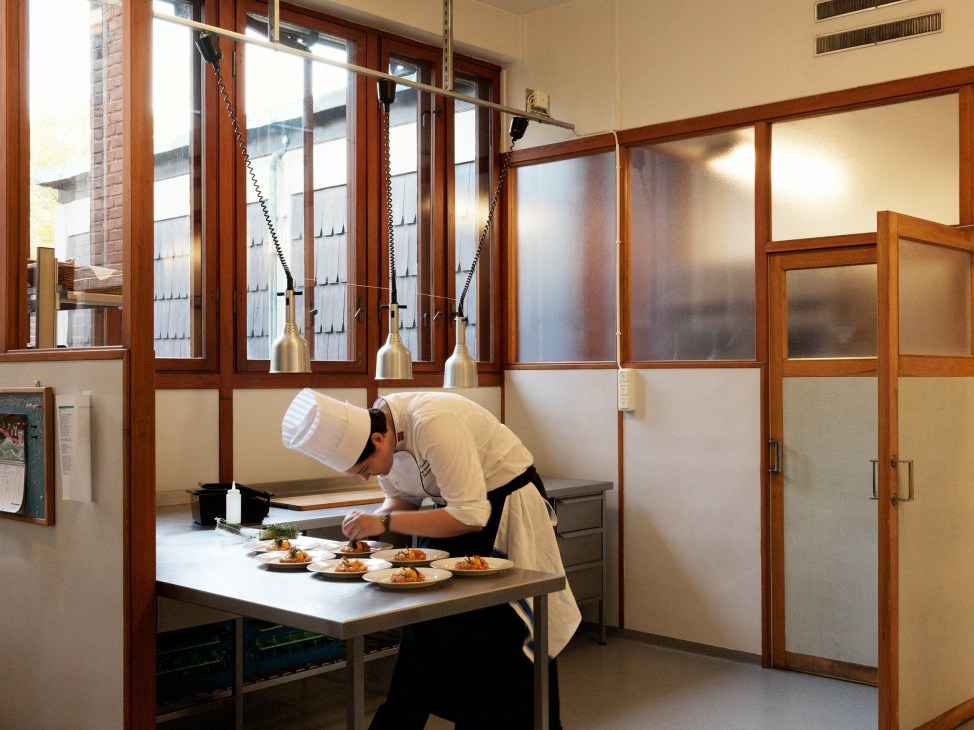 Chef in white chef's coat and hat preparing plated dishes at a counter in a modern kitchen with wooden frames and pendant lights.