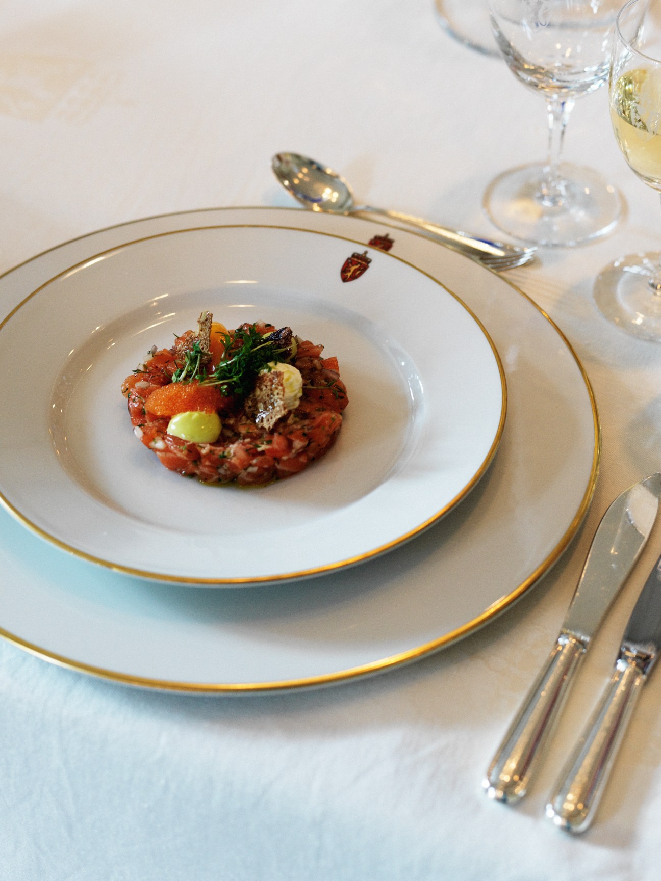Fine dining place setting with Jacob Prytz-designed silver cutlery, white plate with gold rim, and white wine glass.