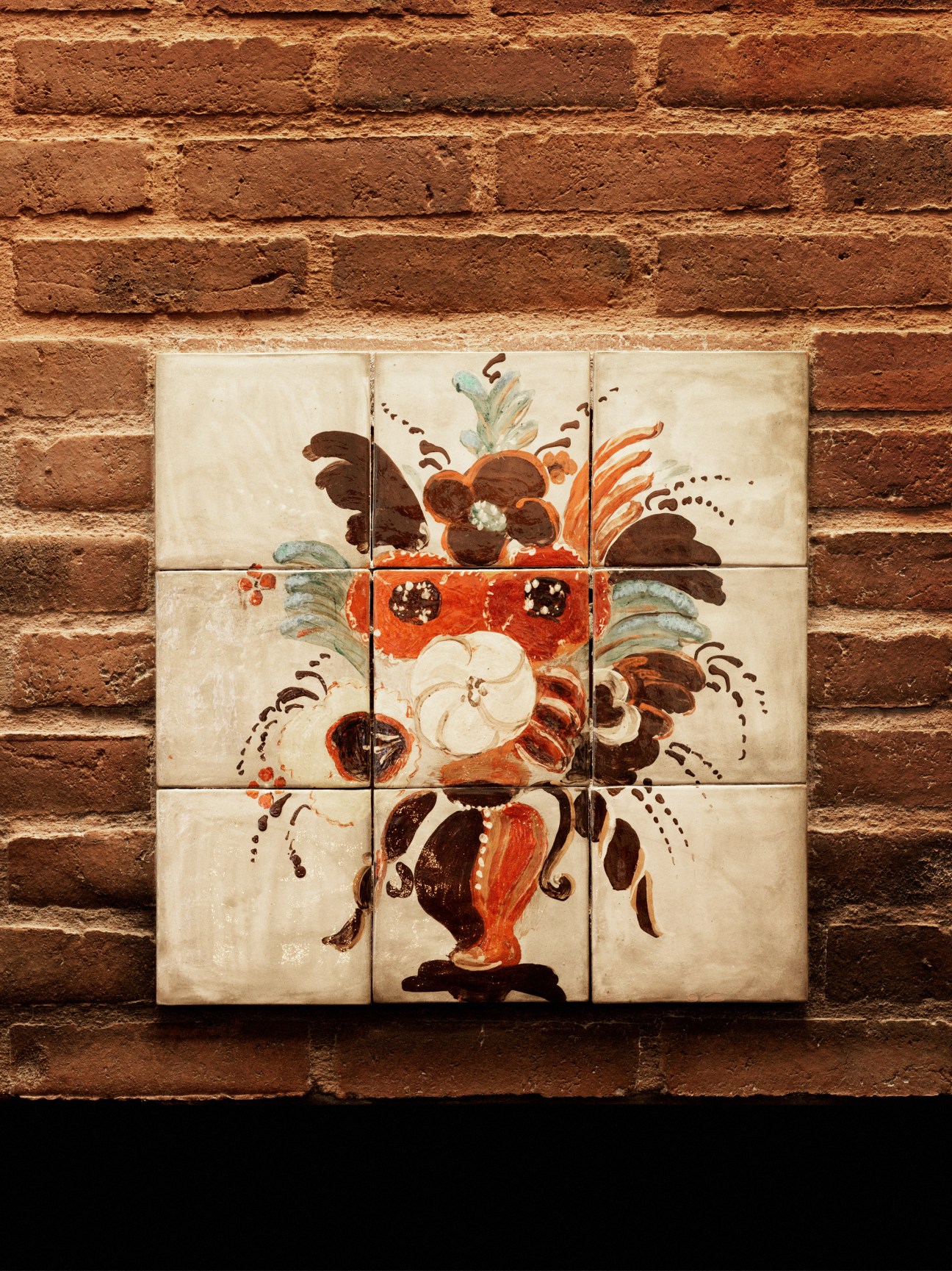 Decorative ceramic tile mural mounted on a brick wall, depicting a classical floral arrangement in a red vase with brown flowers and green foliage.