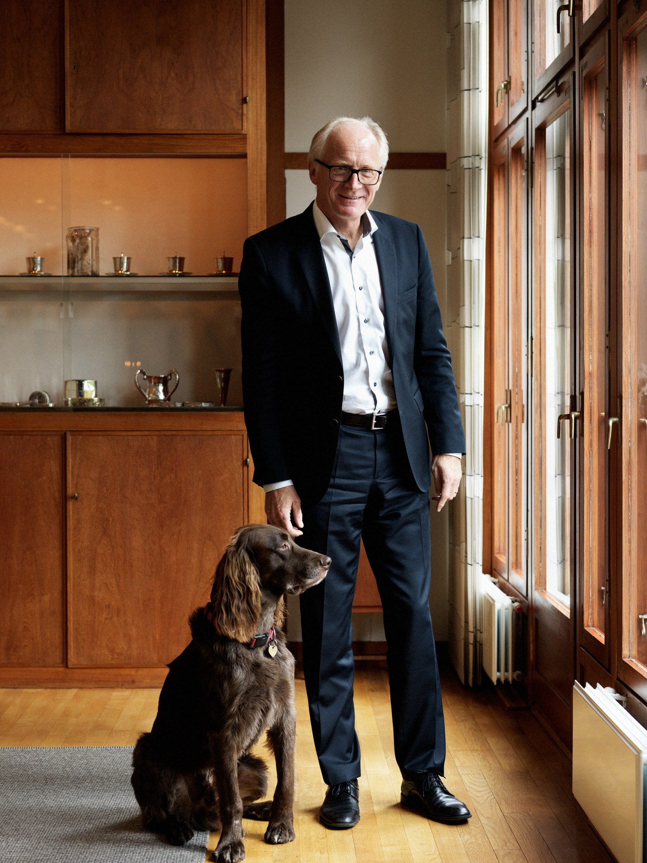 Ambassador Kai Eide standing in a modernist interior with wooden cabinetry and his dog beside him.