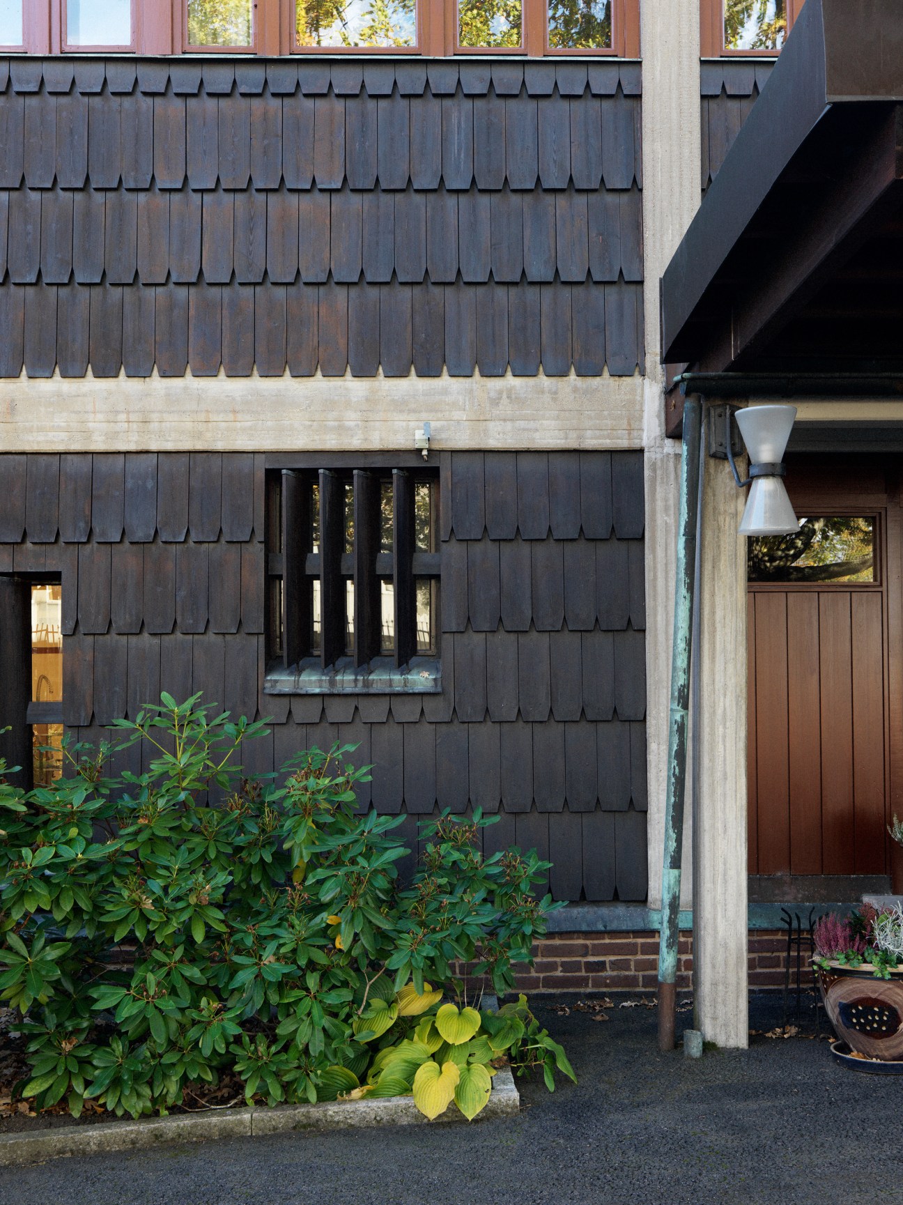 Dark wooden shingle exterior wall of Norwegian embassy with windows and entrance surrounded by green foliage.
