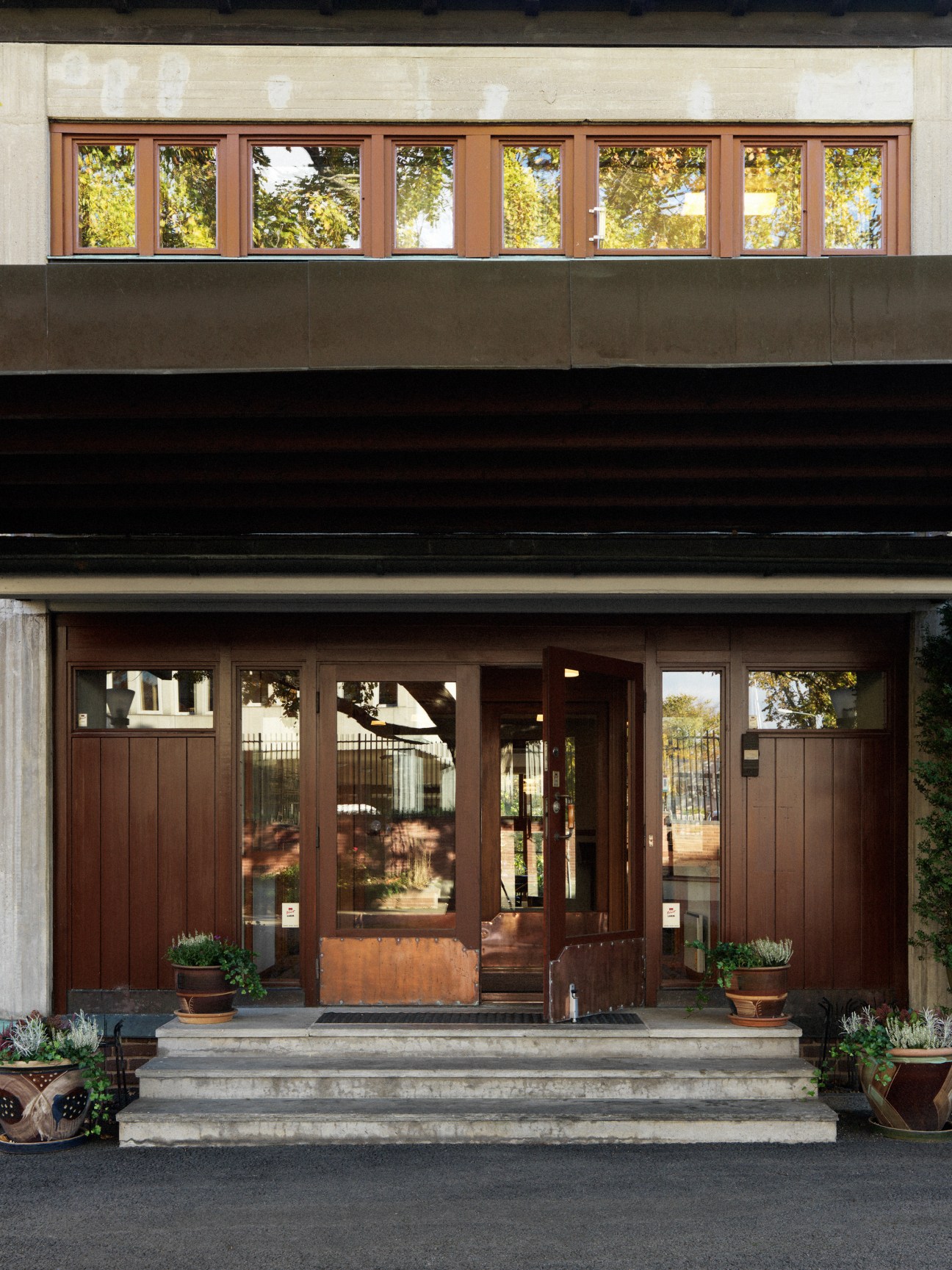 Two-storey modernist building facade with brown wooden doors, windows and stepped concrete entrance flanked by potted plants.