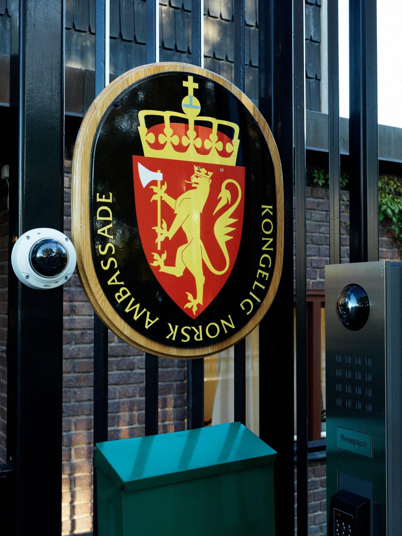 Oval wooden-framed plaque with Norwegian royal coat of arms and text reading 'Ambassade Norsk Dillongkoje' mounted on building entrance.