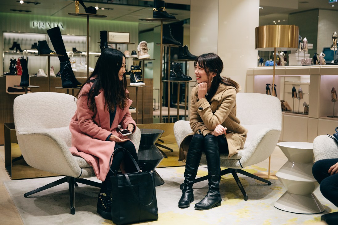 Two women sitting on white chairs in Kadewe's redesigned women's fashion department, with shoe displays and Prada signage visible in the background.