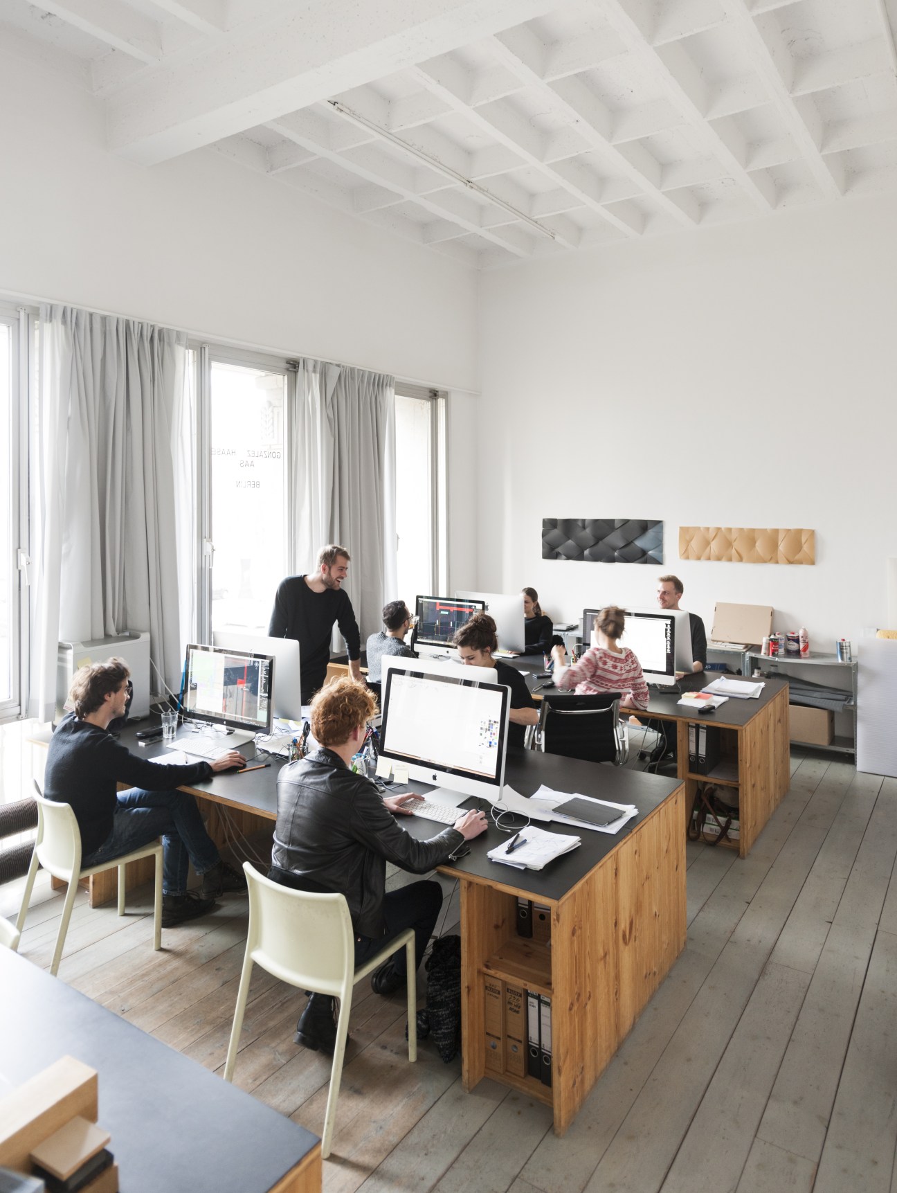 Interior of a bright office space with employees working at computers at wooden desks with white chairs, white coffered ceiling, and large windows with gray curtains.