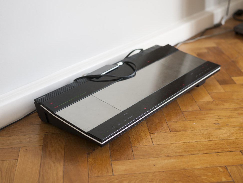 Black stereo receiver with control panel and cables on wooden floor against white baseboard.