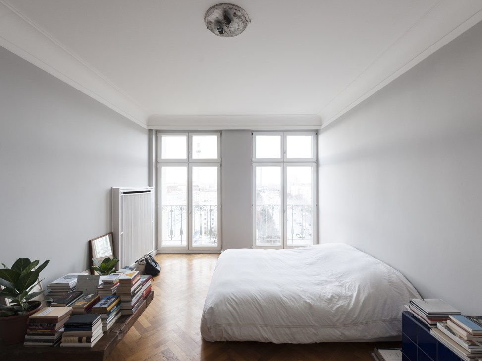 Minimalist bedroom with white walls, large windows overlooking Berlin, wooden floors, and a simple bed with white bedding.
