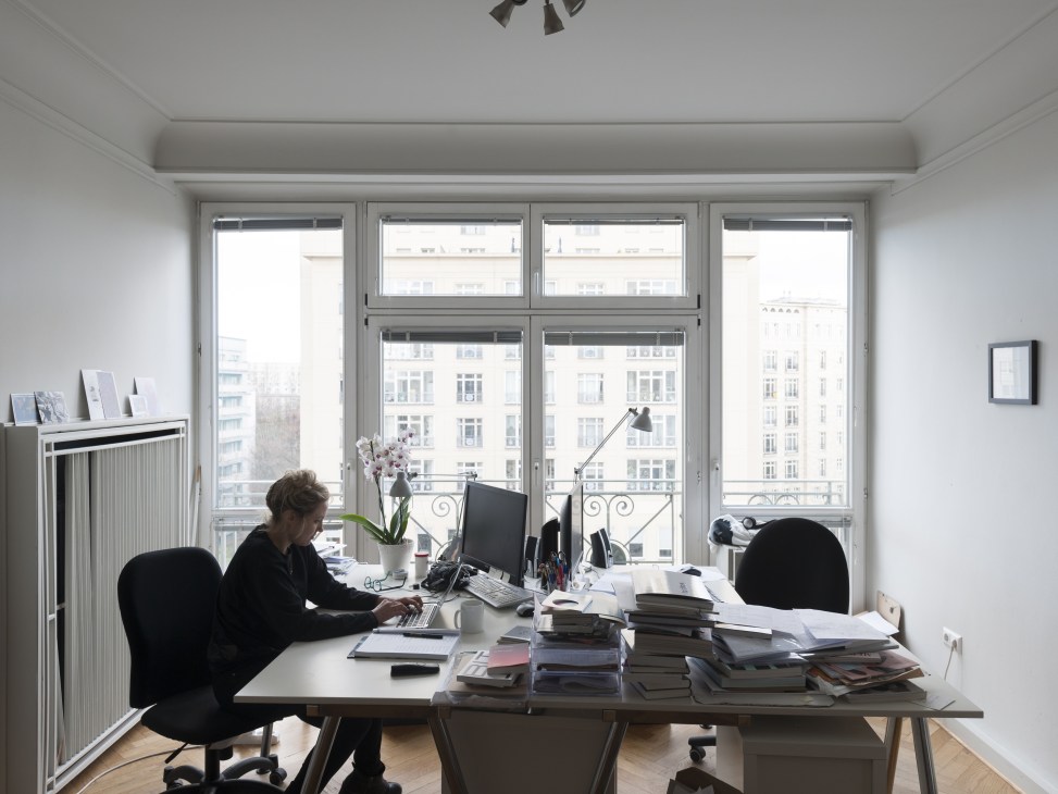 Woman working at desk in bright office with large windows overlooking Berlin apartment buildings.