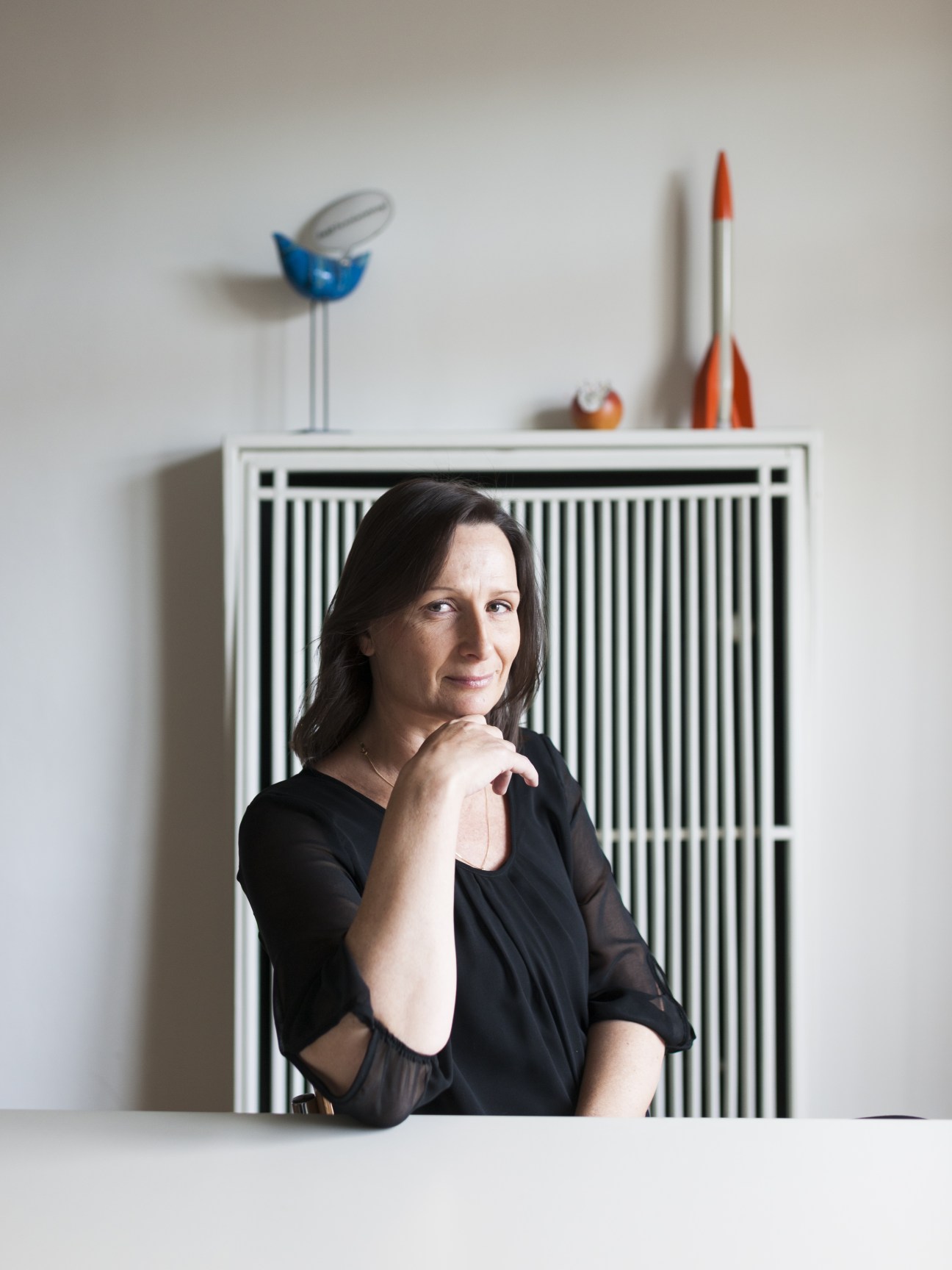 Sabine Ofenbach, managing director of Texte zur Kunst magazine, posing with hand to chin in front of striped wall panel with toy models on shelf.