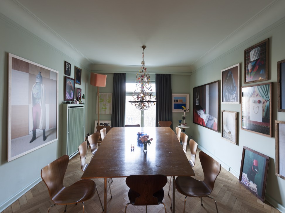 Dining room with mid-century wooden chairs around a long dark table beneath a decorative chandelier, with framed artwork covering mint-green walls.