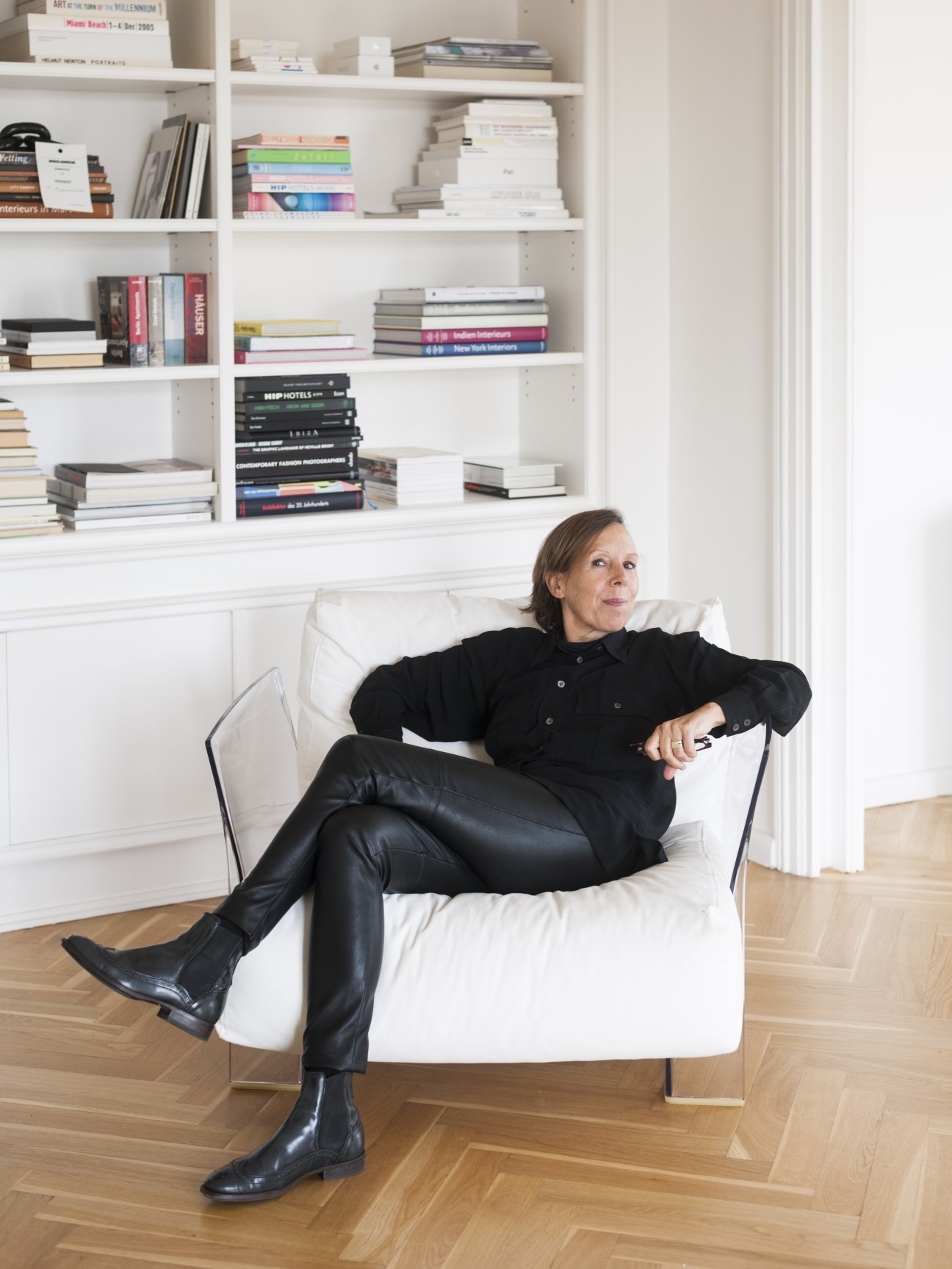Woman in black clothing seated in white chair against shelving unit filled with books in Berlin apartment.