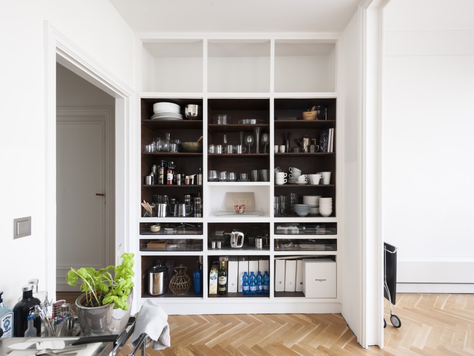 A minimalist white shelving unit displaying tableware, kitchenware, and decorative objects against white walls.