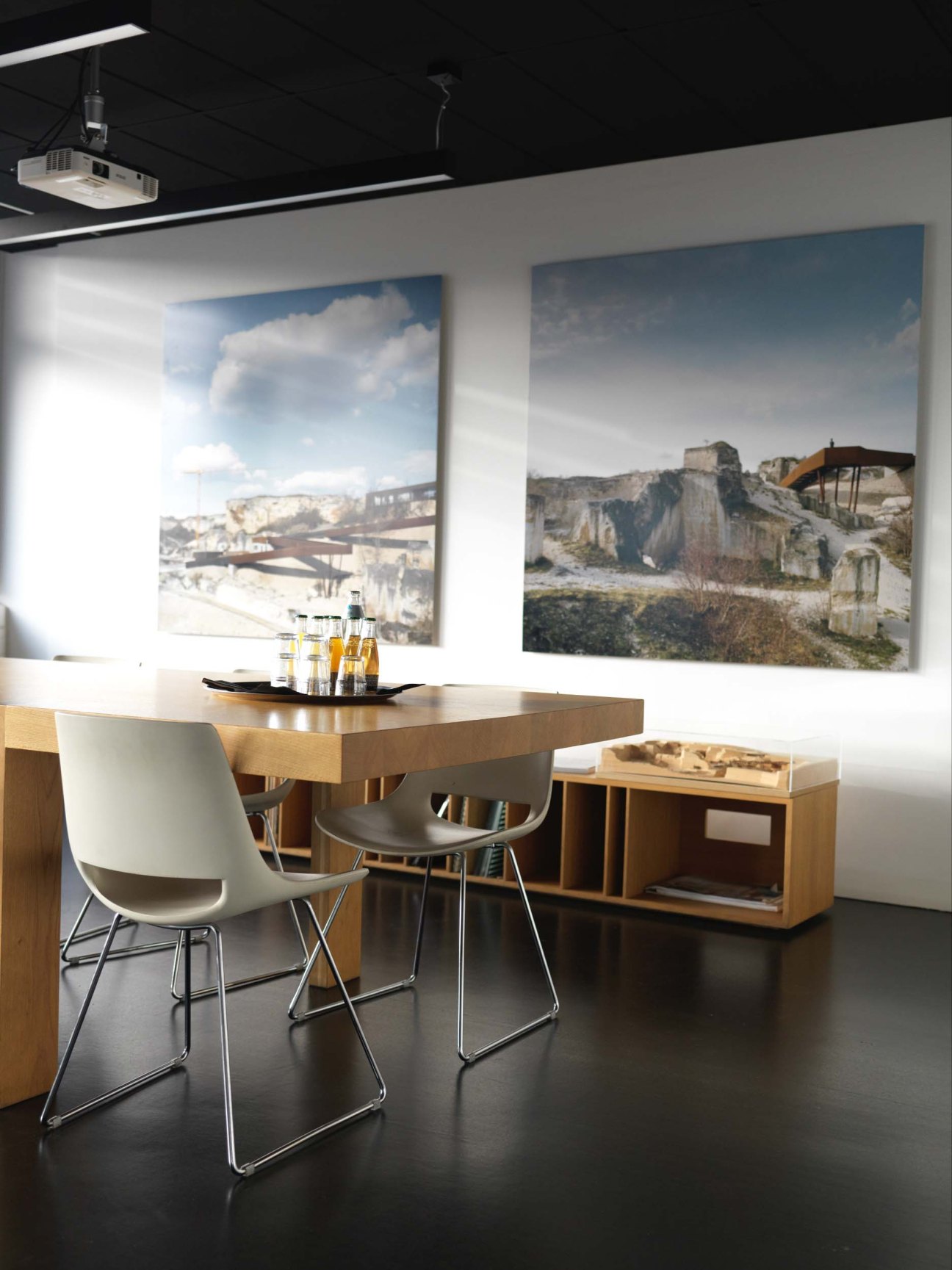 Modern meeting room with wooden desk, white chairs, and large framed photographs of industrial landscapes on white walls.