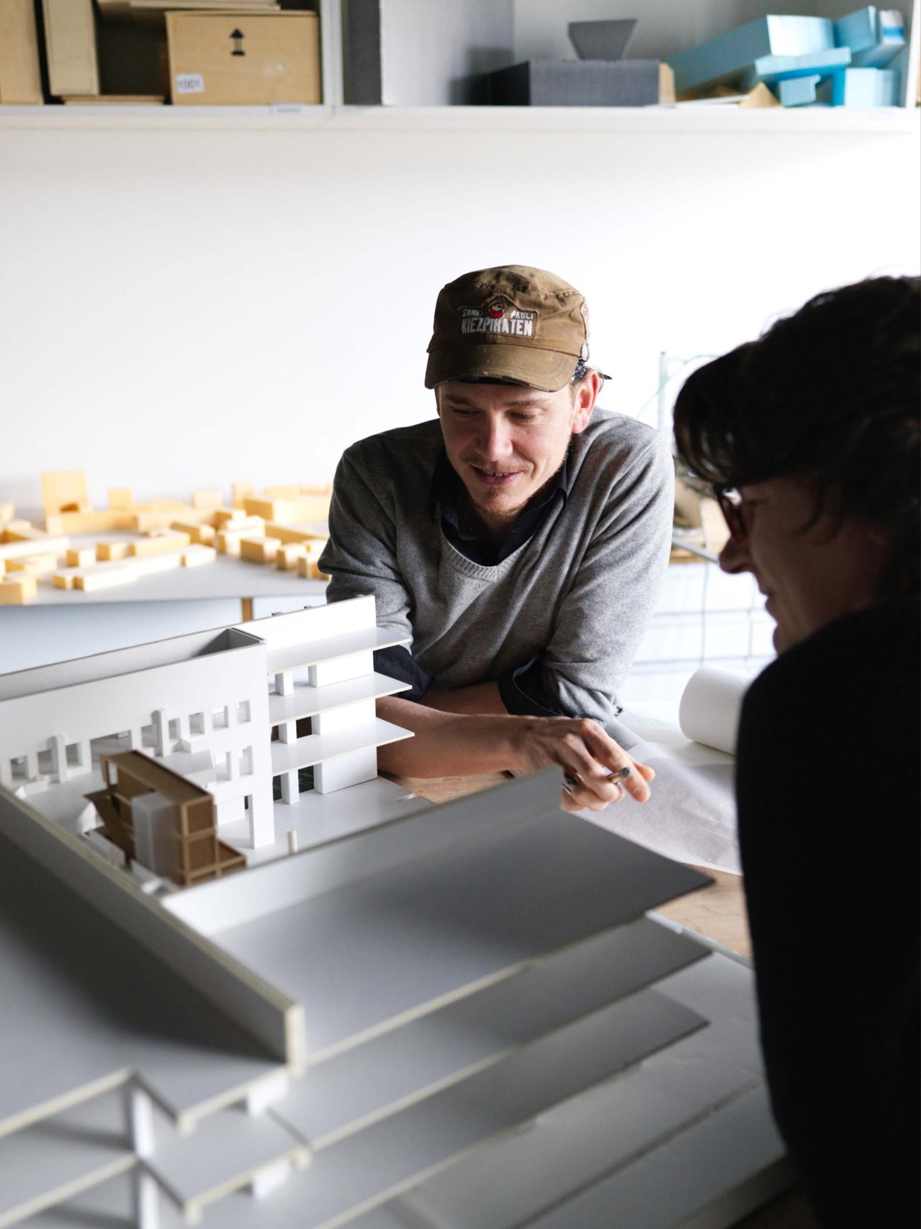 Man in cap smiling while discussing architectural models with another person at a desk.
