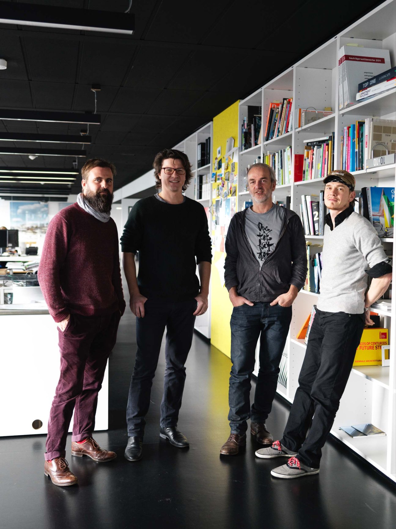 Four men standing in a modern office with white shelving and yellow accent wall behind them.