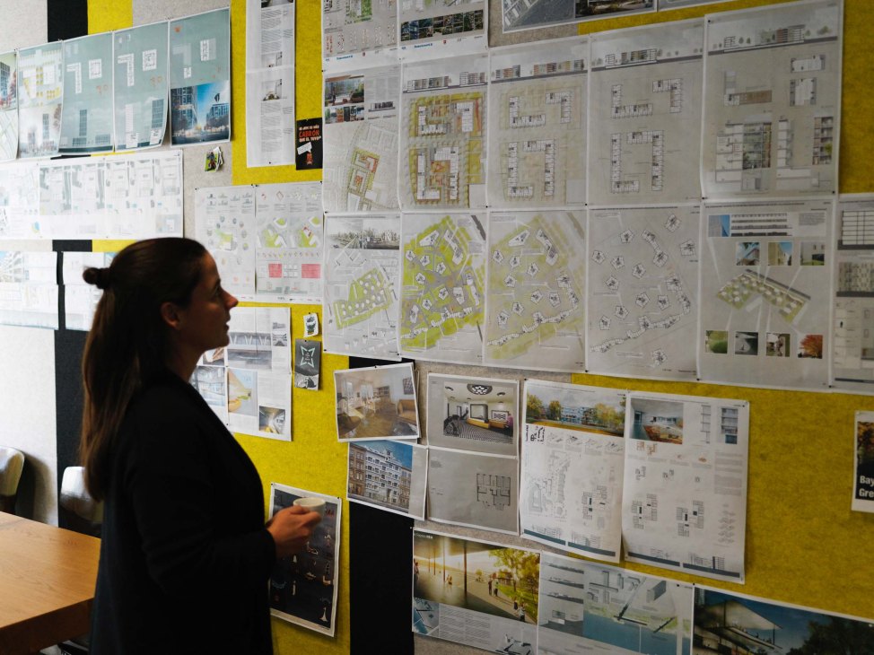 Woman viewing architectural plans and project boards displayed on a yellow wall in an office space.
