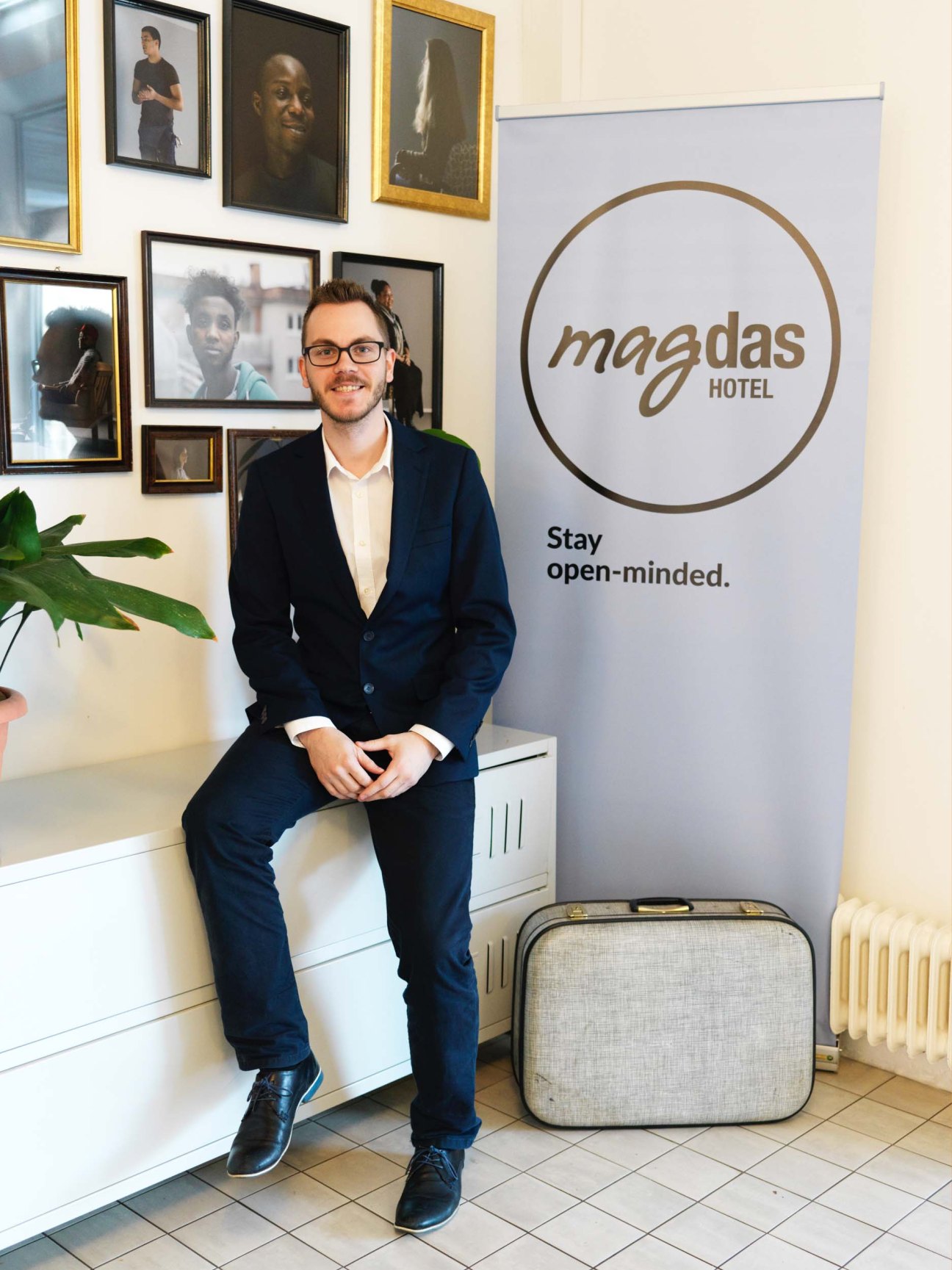 Man in navy blazer sitting in Magdas Hotel lobby with staff portraits and hotel signage behind him.
