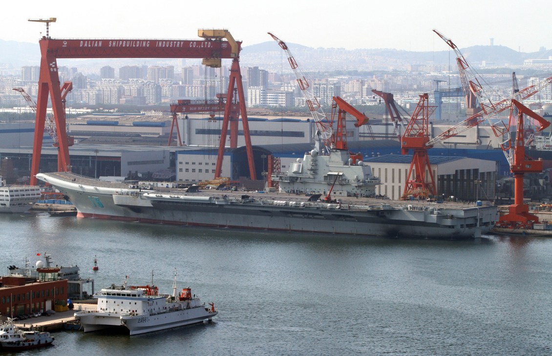 Chinese aircraft carrier under construction at Dalian Shipbuilding Industry shipyard with red cranes and cityscape.