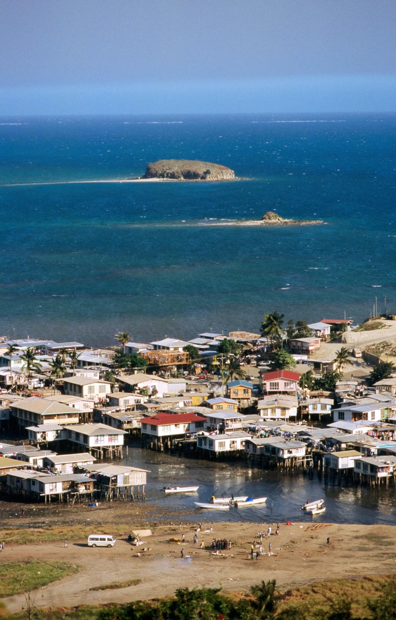 Coastal town in Papua New Guinea with colorful houses built over water, palm trees, and rocky islands visible offshore.