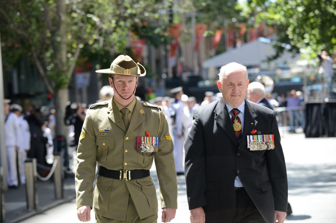 An Australian Defence Force soldier in uniform walks alongside an older man in civilian formal attire wearing military medals during a public ceremony.