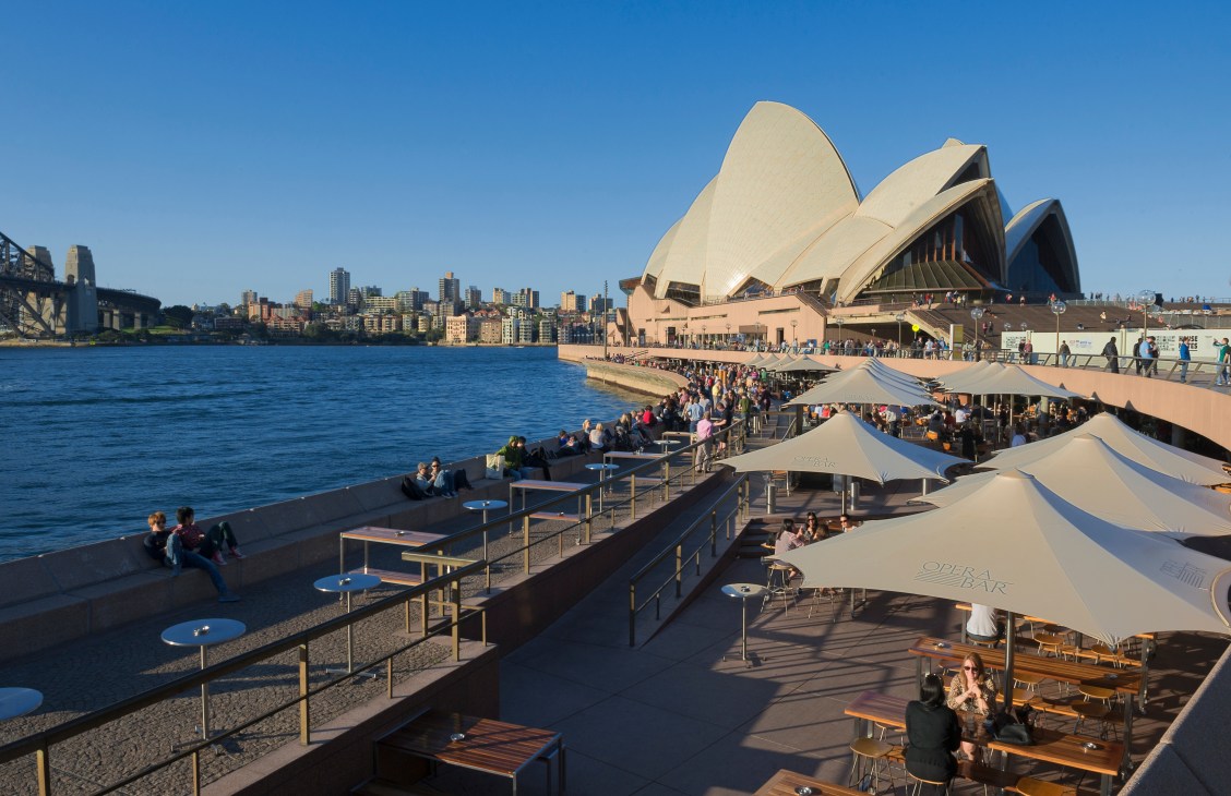 Sydney Opera House and waterfront with crowds of people, cafes with umbrellas, and city skyline.