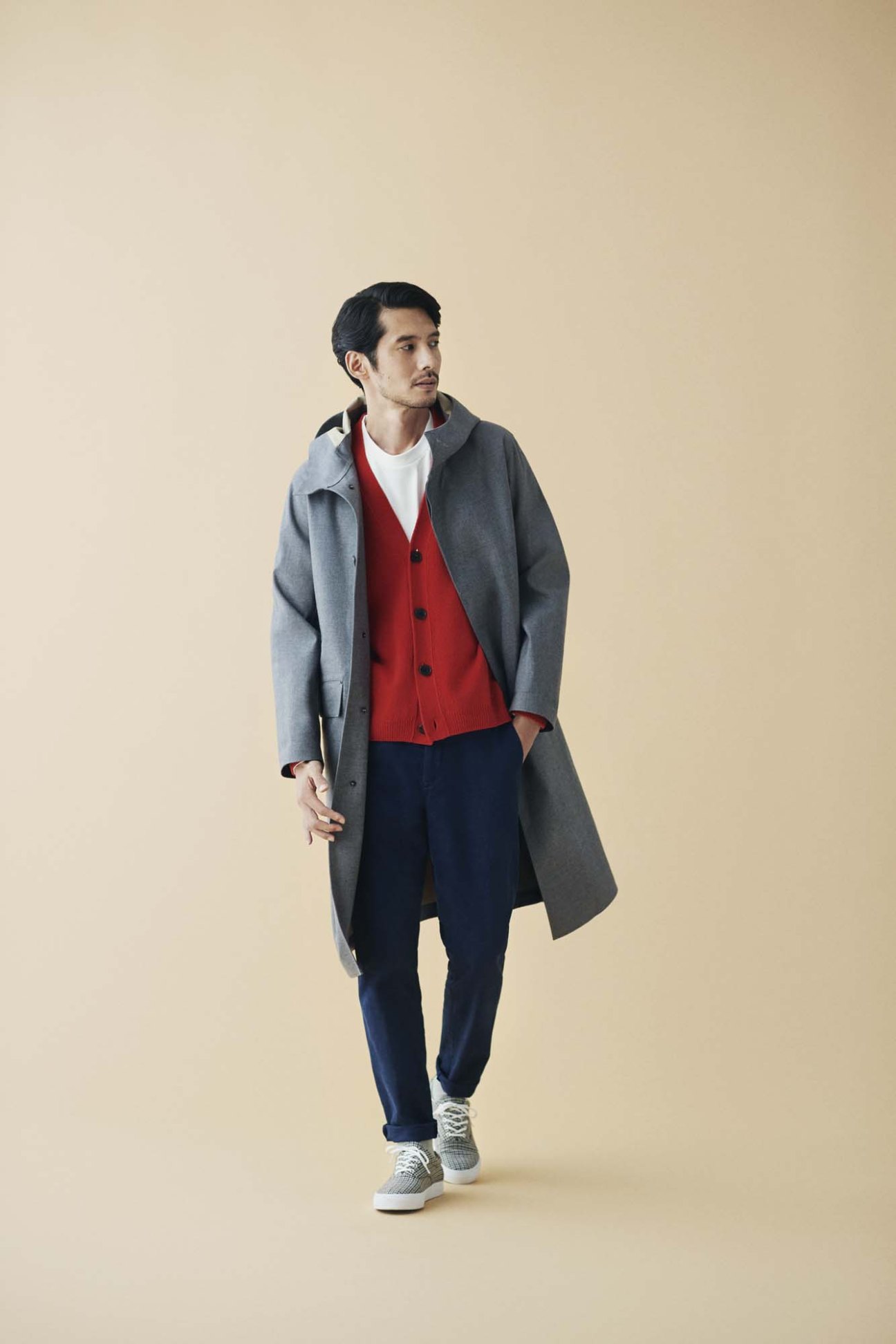 Man wearing gray overcoat, red cardigan, white t-shirt, navy trousers and white sneakers against beige background