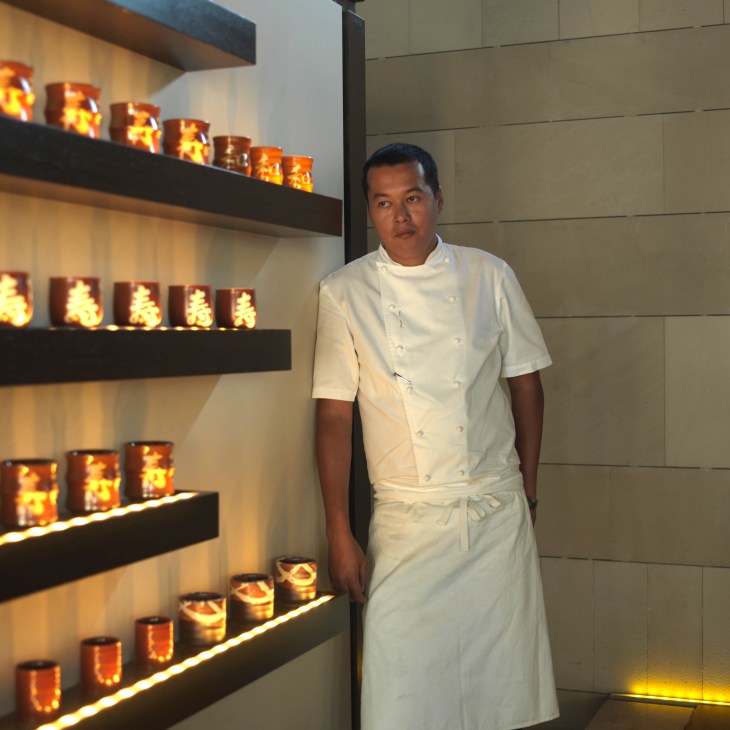 Anthony Sousa Tam, head chef at Chinar restaurant, stands in white chef's whites next to illuminated shelving.