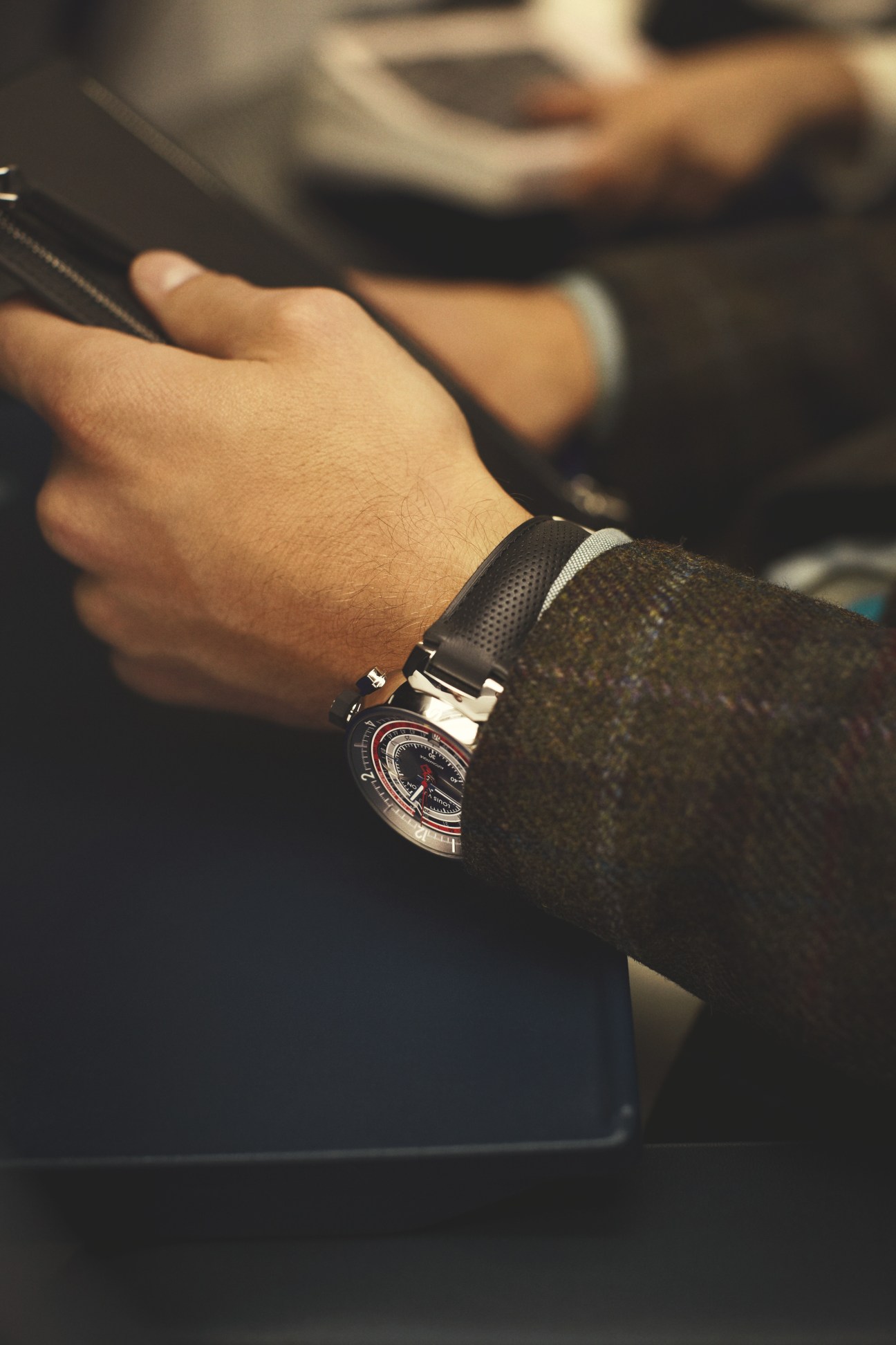 Man's wrist wearing a luxury watch with dark jacket and bracelet, holding a phone.