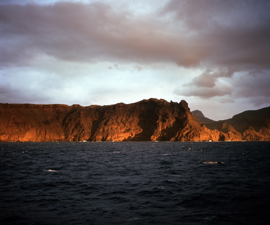 St Helena island's dramatic rocky cliffs illuminated by golden sunlight as the RMS ship approaches from the ocean.