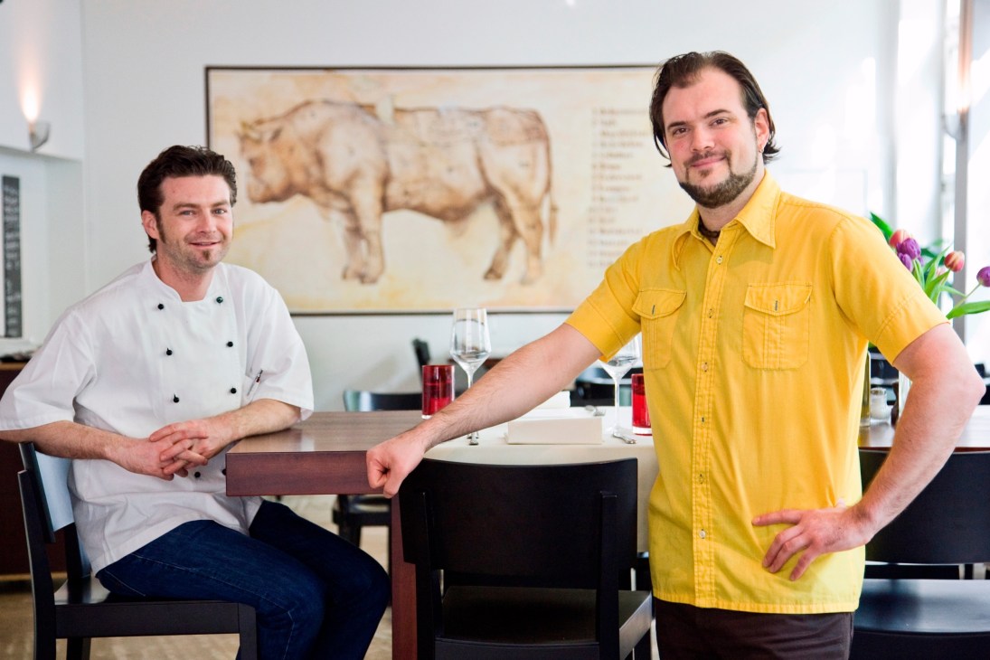 Two men in a restaurant with a cow diagram on the wall behind them.