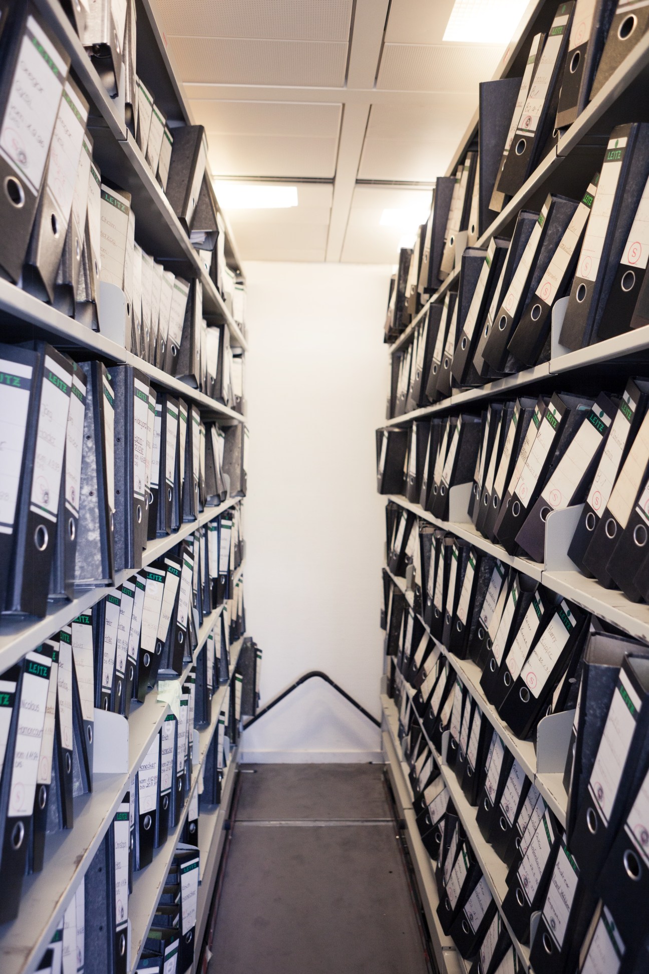 Narrow corridor lined with metal shelving units containing black archive folders and binders labeled with document identifiers.