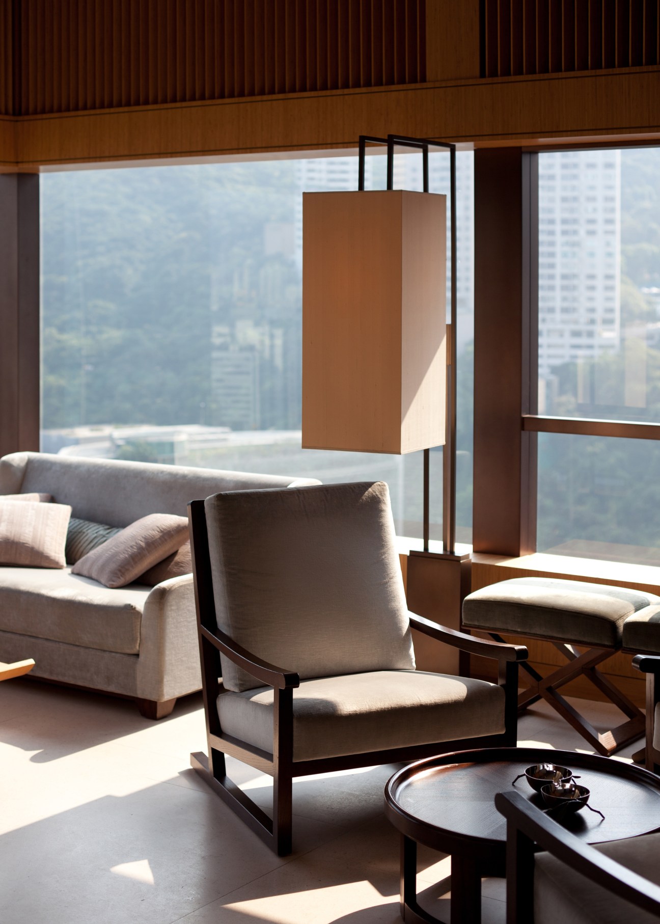 Modern hotel room with floor-to-ceiling windows overlooking a city skyline, featuring minimalist furniture and warm wood tones.