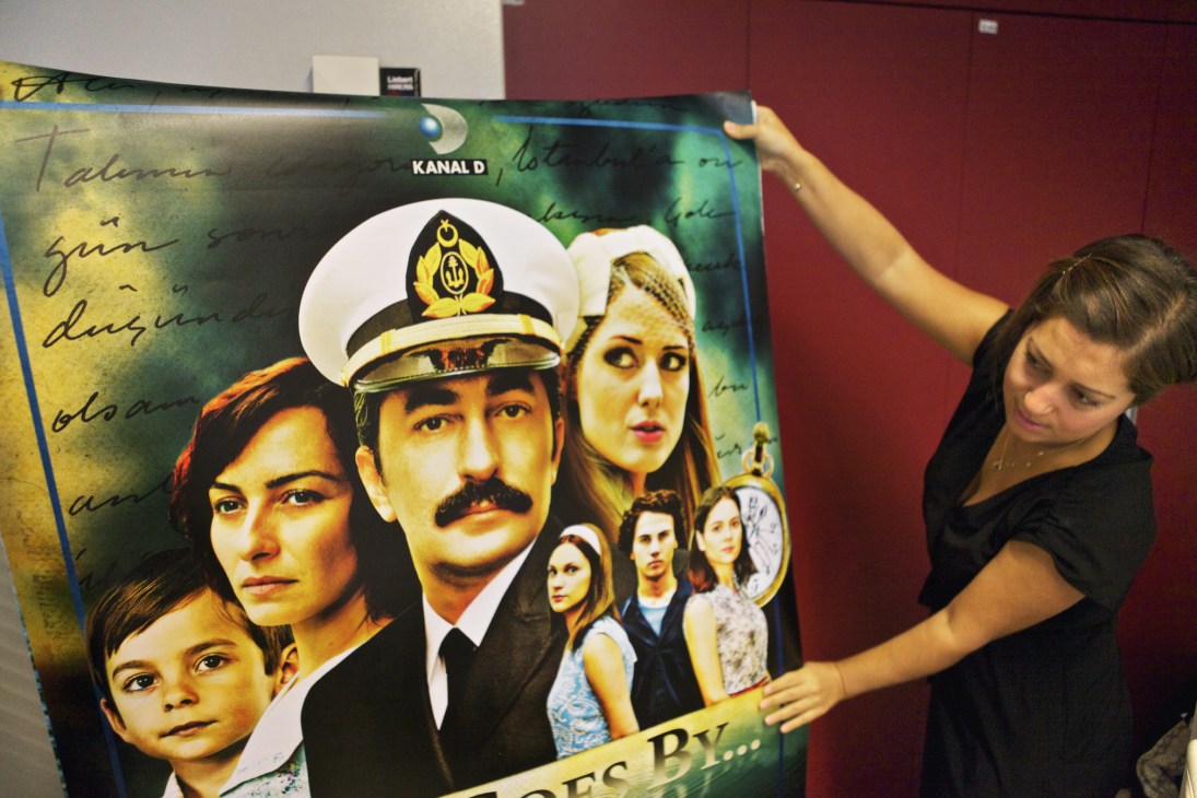 Woman holding a Kanal D poster featuring Turkish drama series cast members and characters in the sales office.