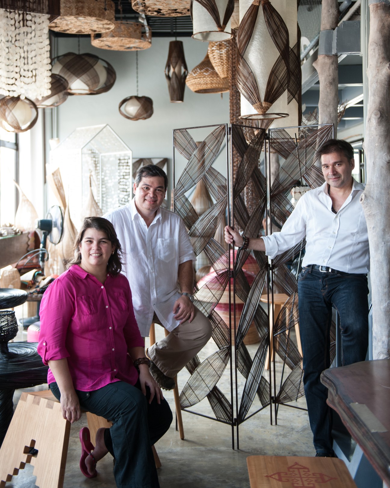 A woman in a magenta shirt and two men in white shirts stand in a showroom with woven baskets and pendant lights displayed overhead.