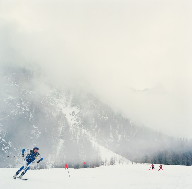 Ski mountaineer in blue outfit racing uphill on snowy slope with misty forested mountains and competitors in background.